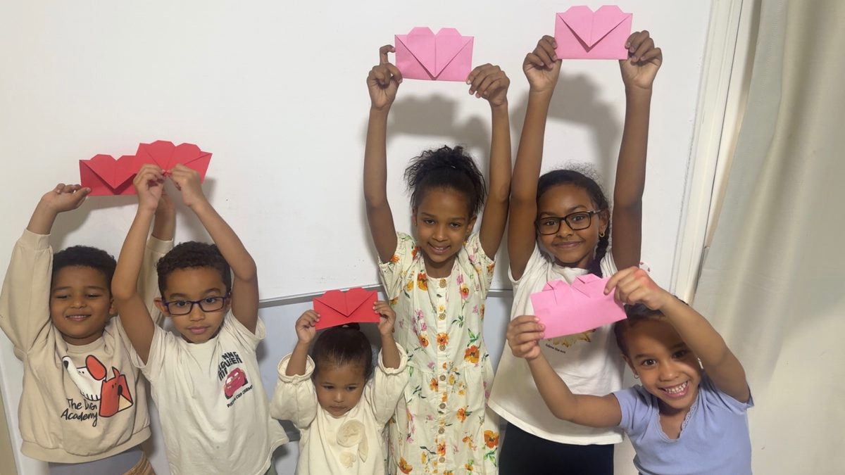 Seven children in a classroom hold red and pink heart envelopes over their heads, smiling.