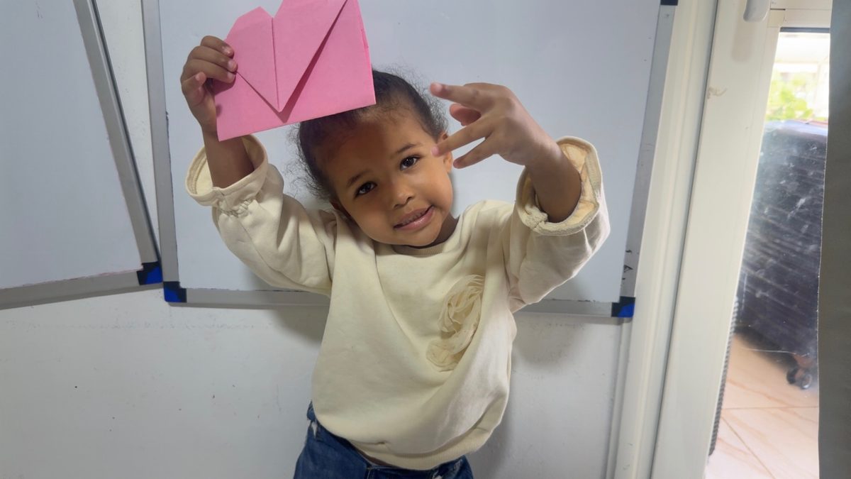 A toddler holds a red heart envelope over her head while an older girl beside her raises hers.