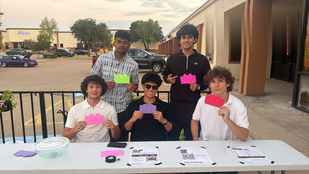 Five team members at a folding table with flyers, a donation bowl, and pink, green, and purple envelopes, a parking lot and cars behind them at dusk.