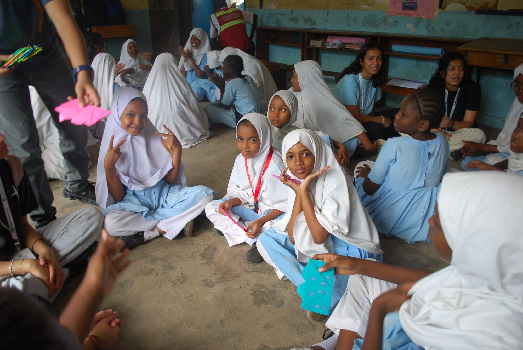 A Grade 4 class at Central Girls Primary School sits in a circle on the classroom floor as a volunteer hands out a pink HeartPack envelope; two girls flash peace signs at the camera and another holds a teal envelope.