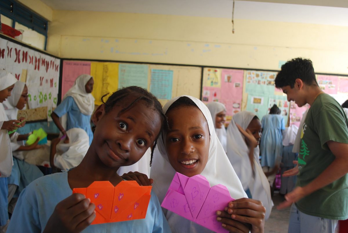 Two girls stand shoulder to shoulder holding envelopes covered in hand-drawn hearts, one beaming at the camera.