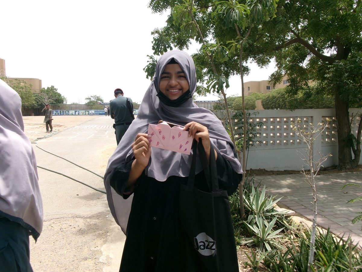 A girl in a grey hijab laughs while holding a pink envelope decorated with hand-drawn hearts.